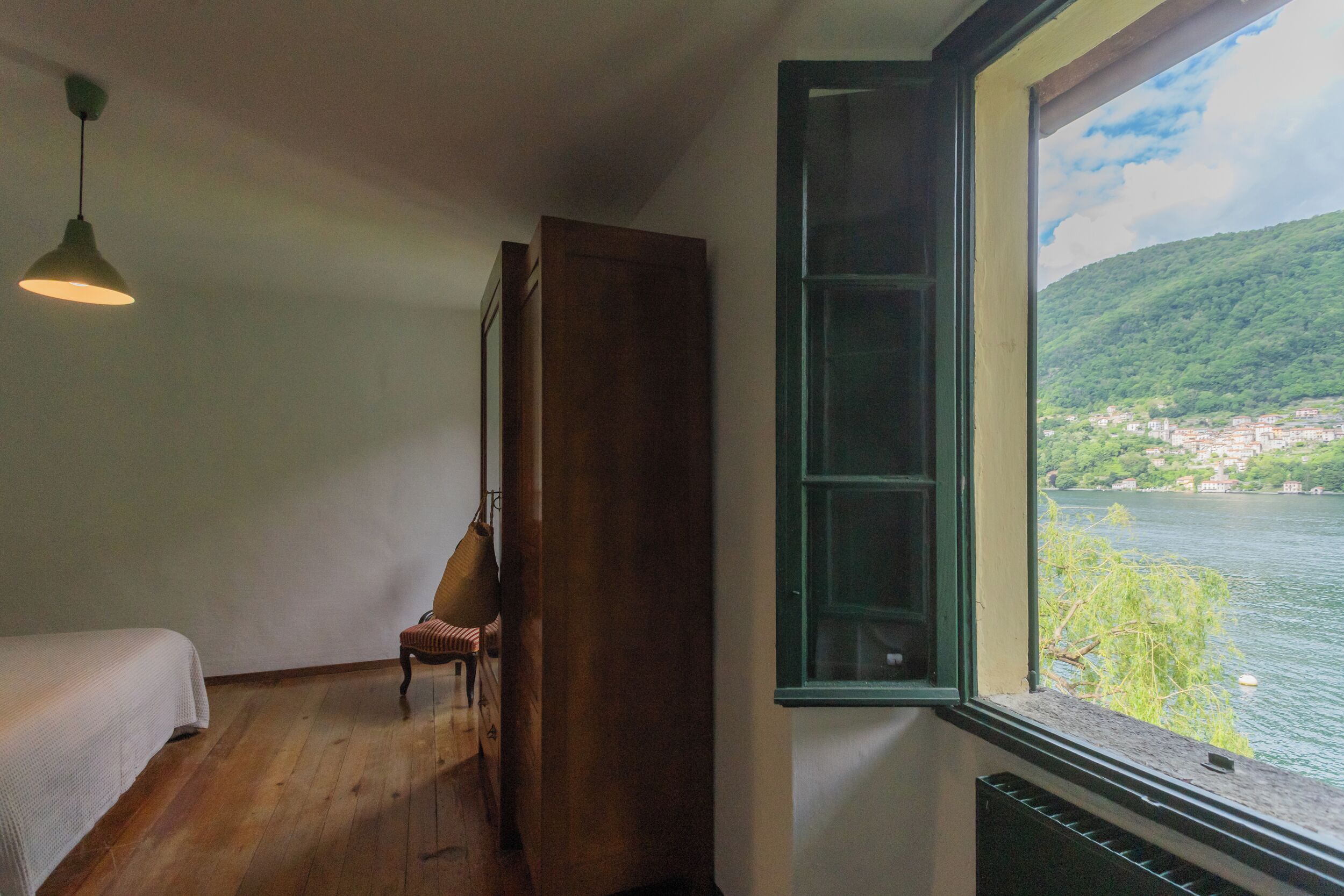 A room with a bed, a chair, a window with a view of a lake and a town, and a hanging light fixture.