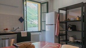 A kitchen with a window view of a green hillside, a refrigerator, a sink, and a wooden cutting board with a loaf of bread and an open book. - Residenza Regina by Rent all Como (Laglio)