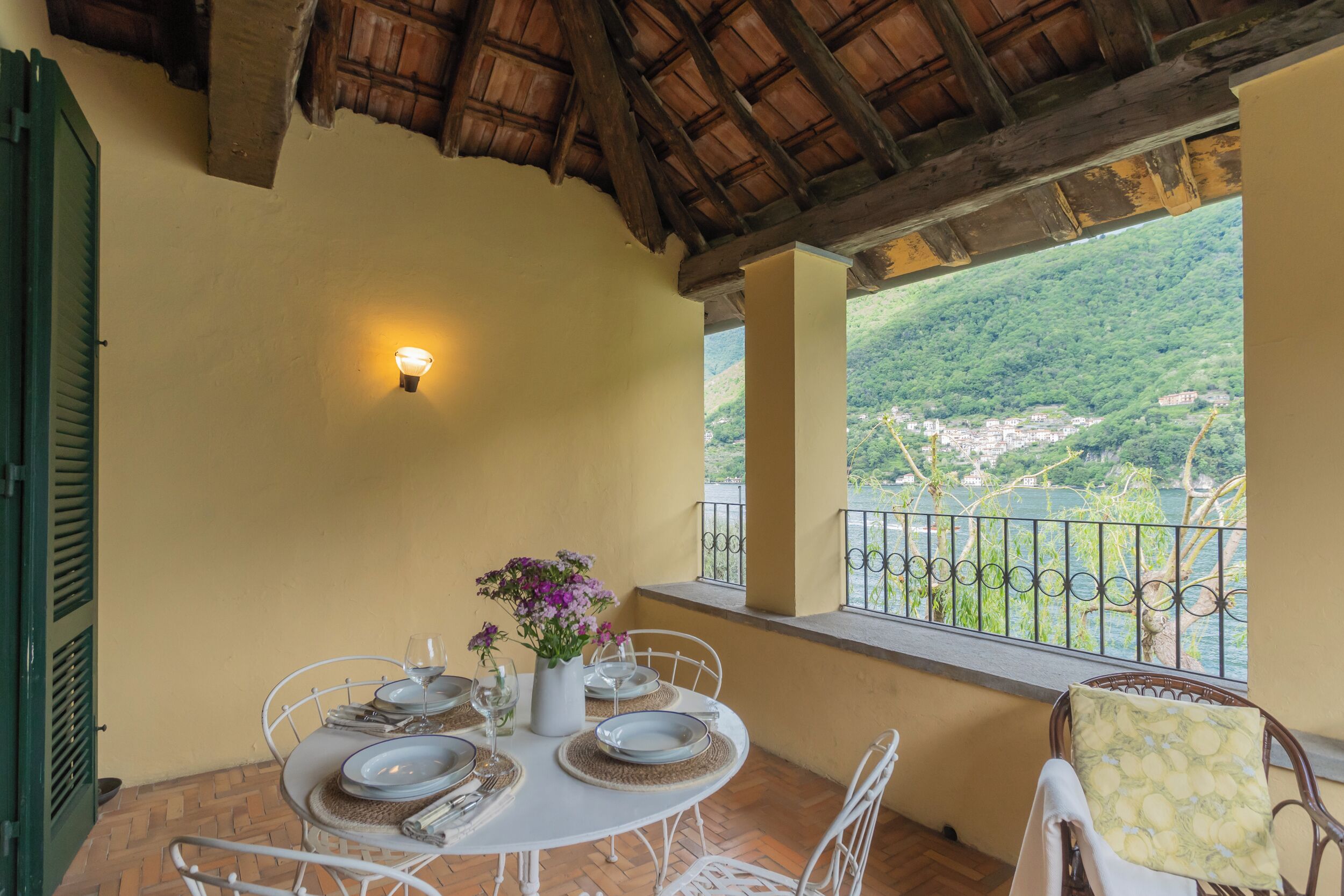 A dining area with a table set for four, a vase with flowers, and a view of a mountainous landscape through a balcony railing.