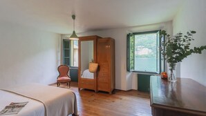 A bedroom with a bed, a chair, a wooden wardrobe, a window with a view, and a hanging light fixture. - Residenza Regina by Rent all Como (Laglio)