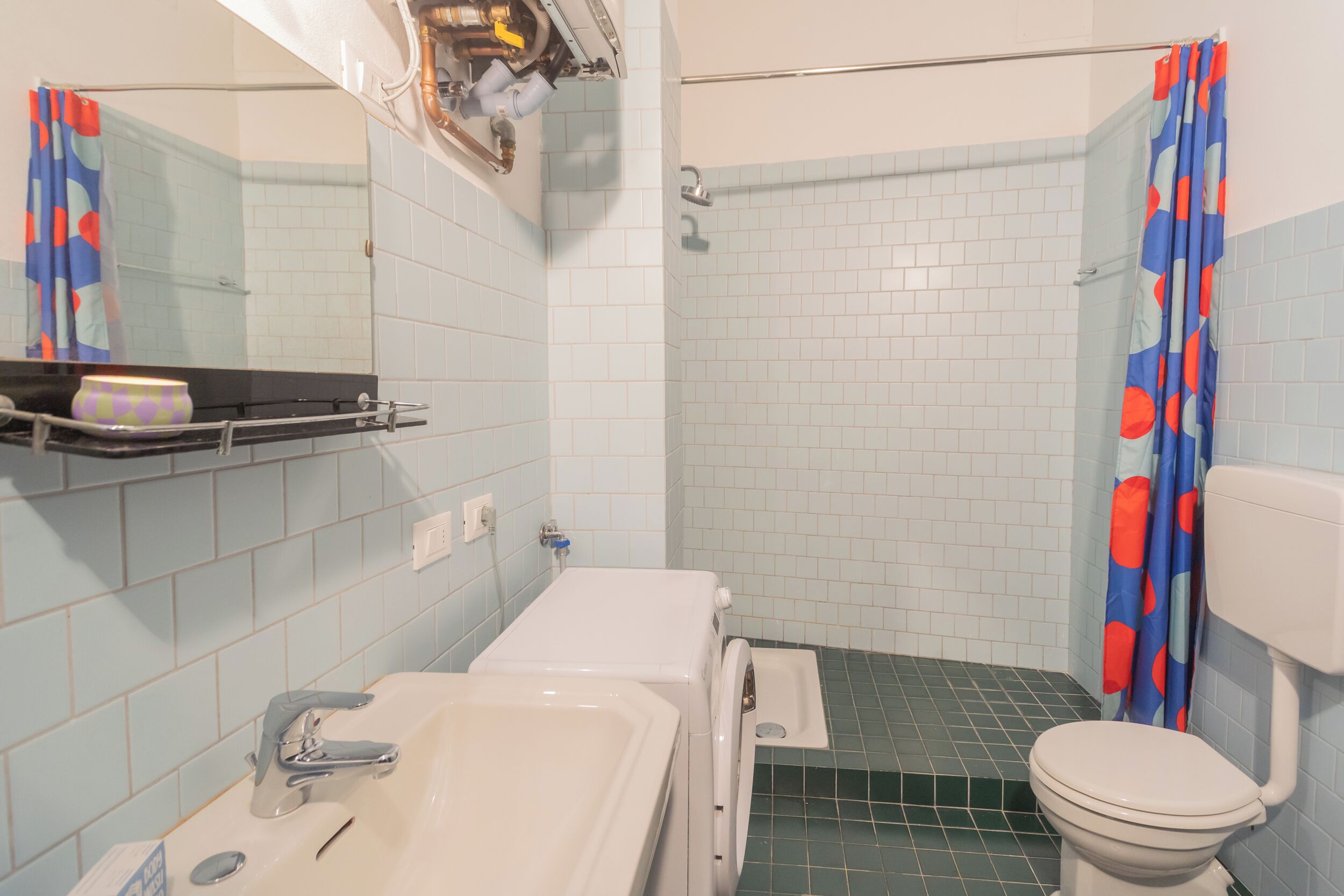 Une salle de bain avec un lavabo blanc, des toilettes blanches, une douche avec un rideau bleu et rouge, et des murs recouverts de carreaux bleu clair.