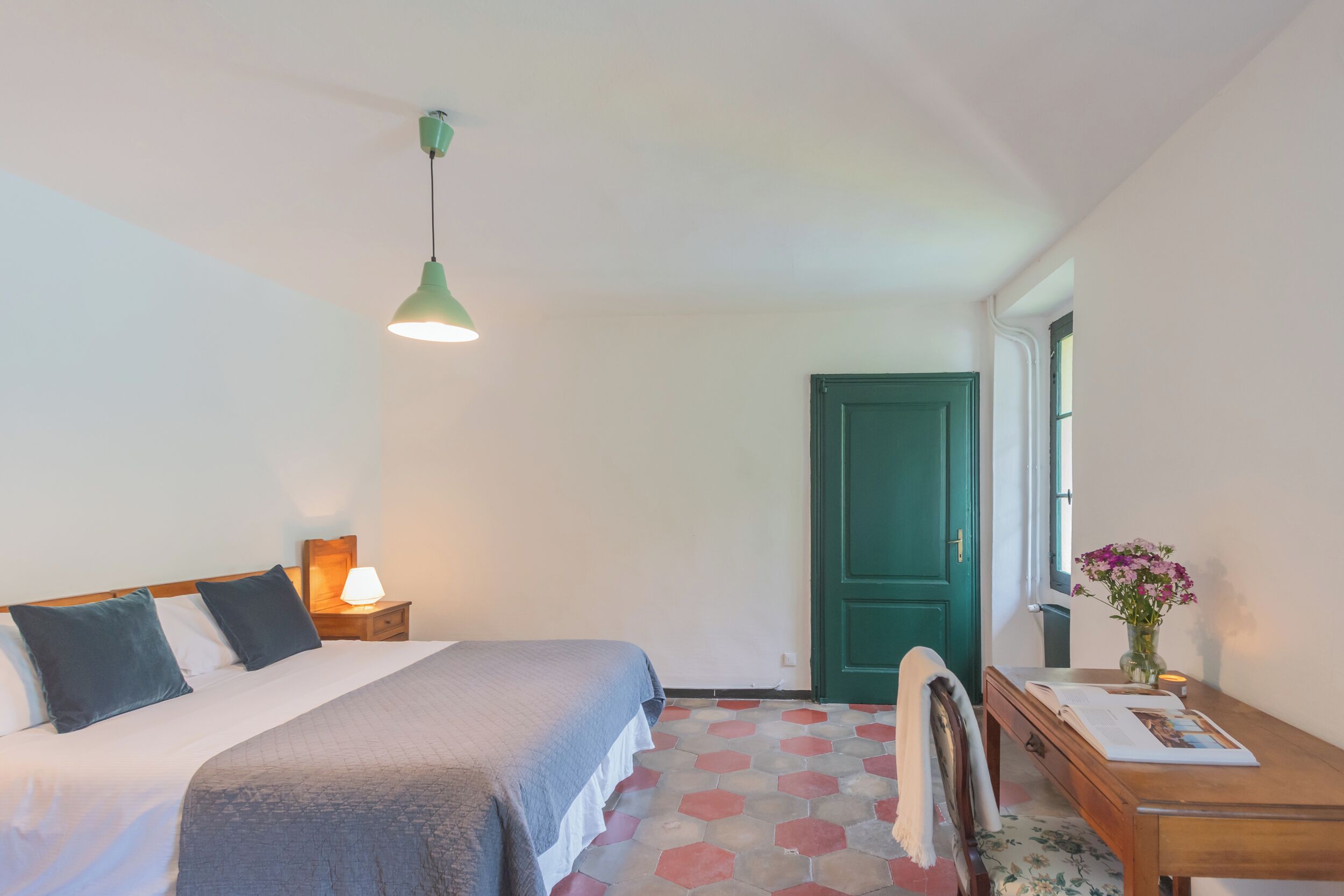Ein Schlafzimmer mit einem Bett, einem Nachttisch mit Lampe, einem Holztisch mit Buch und Blumen sowie einer grünen Tür.