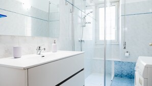 A modern bathroom with a glass shower enclosure, white marble tiles, and a built-in washing machine.