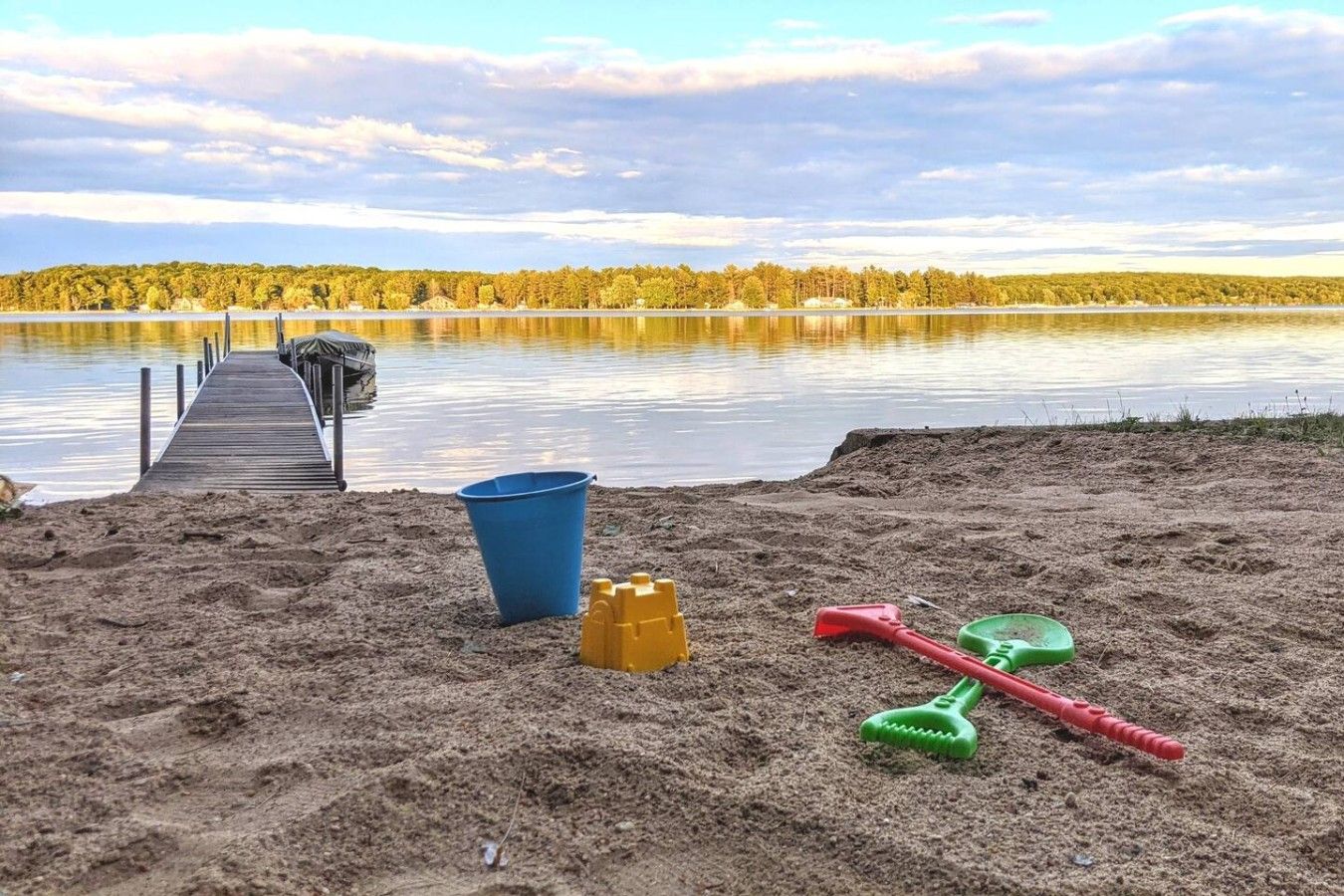 Une plage de sable avec une jetée en bois qui s’avance sur un lac paisible, le tout entouré d’arbres.