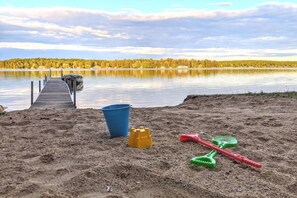 A sandy beach with a wooden dock extending into a calm lake, surrounded by trees.