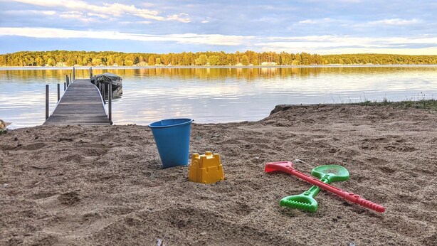 En sandstrand med en träbrygga som sträcker sig ut i en lugn sjö, omgiven av träd.