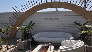 Un espace extérieur lumineux, aménagé pour se détendre, avec un canapé blanc et deux chaises blanches, une petite table, et une arche décorative en bois. Le mot « CINNette » est inscrit sur le mur situé derrière.