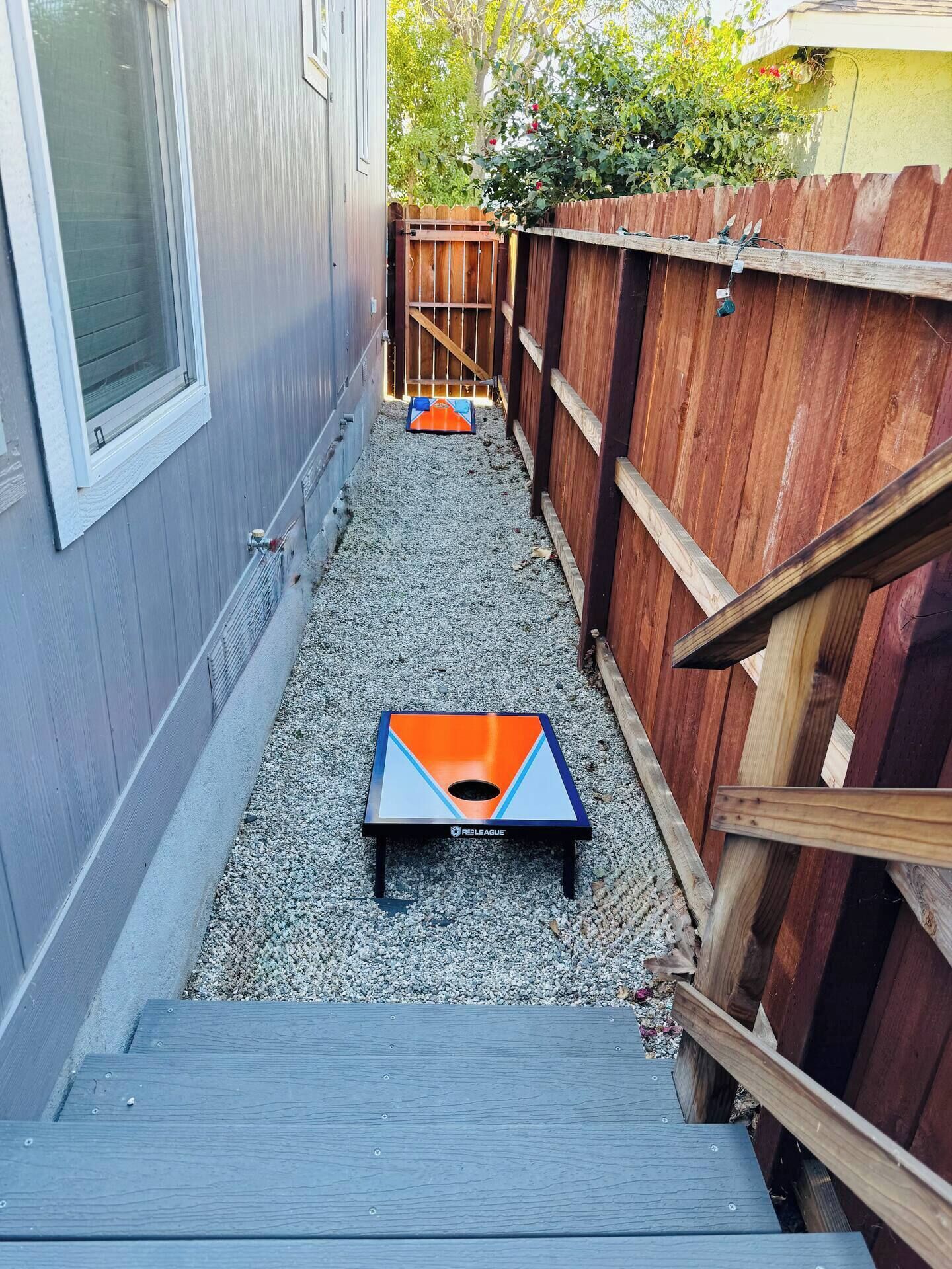 A narrow gravel pathway leading to a small, colorful disc golf basket labeled "RELEAGUE" in a residential backyard with a wooden fence and a house in the background.