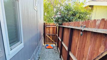 A narrow gravel pathway leading to a small, colorful disc golf basket labeled "RELEAGUE" in a residential backyard with a wooden fence and a house in the background.