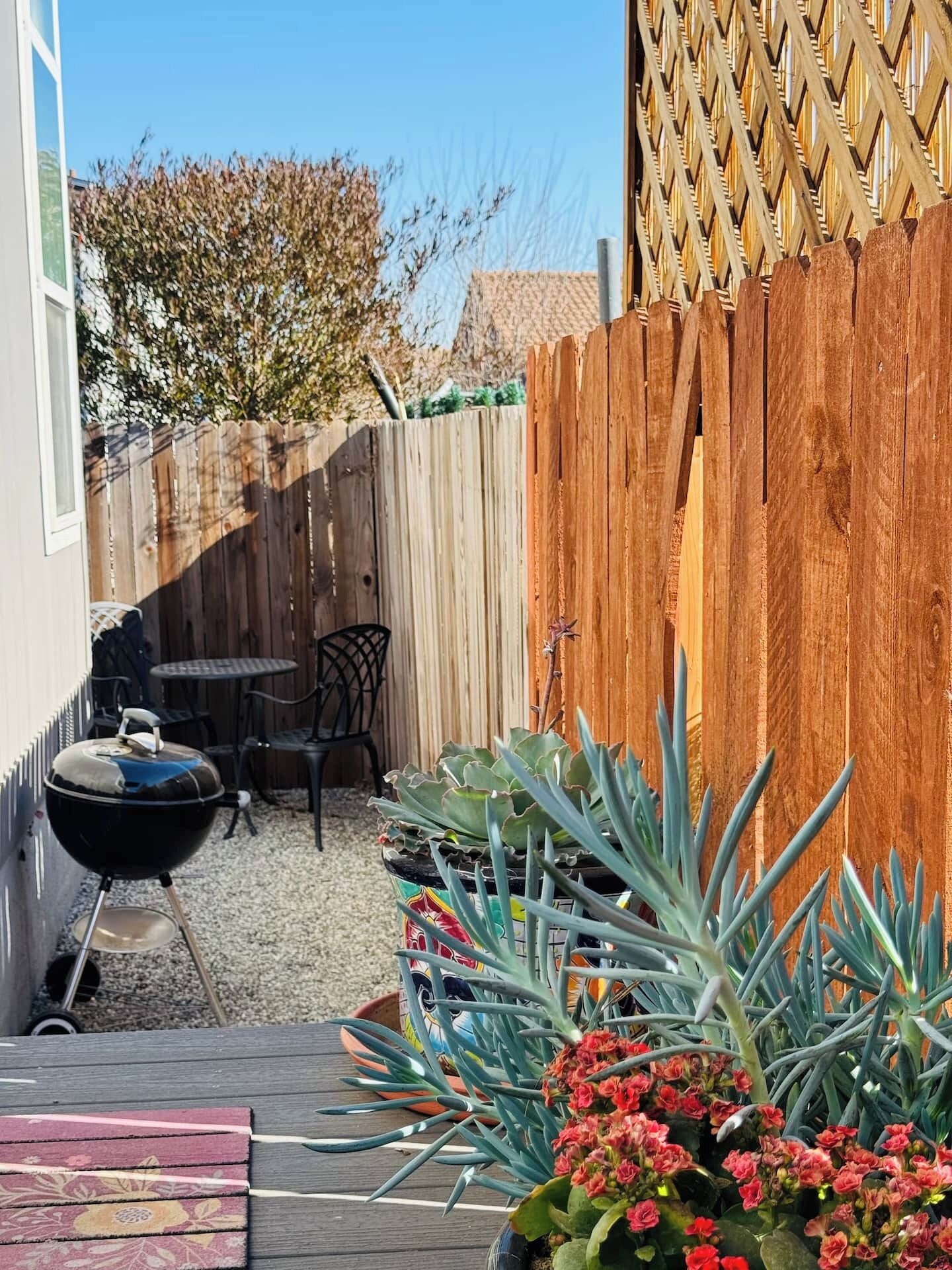 A backyard with a barbecue grill, a wooden fence, a table, chairs, and various potted plants.