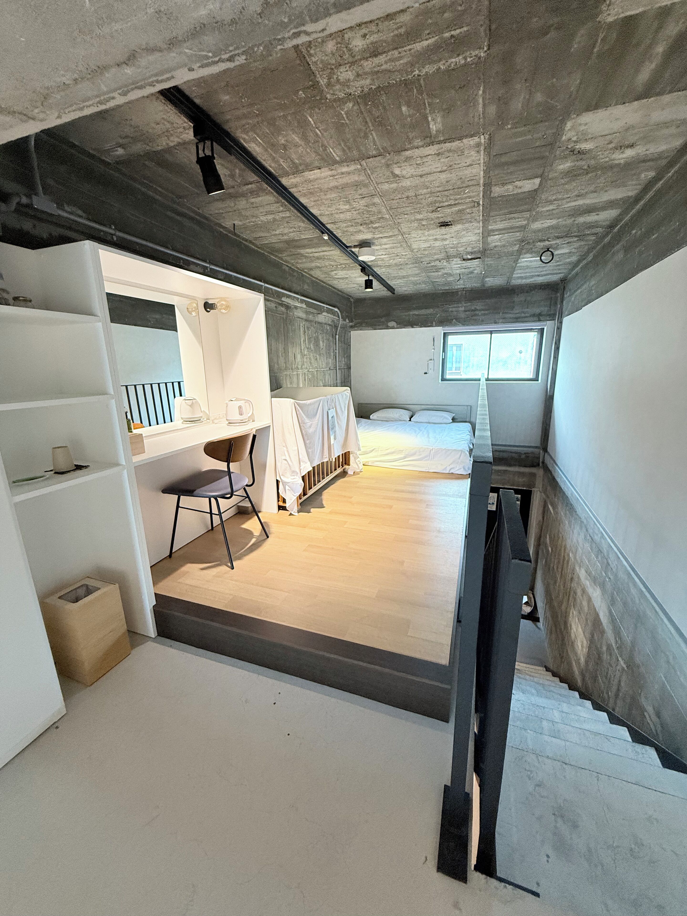 A compact, modern bedroom with a bunk bed, a desk with a chair, a large window, and a concrete ceiling.