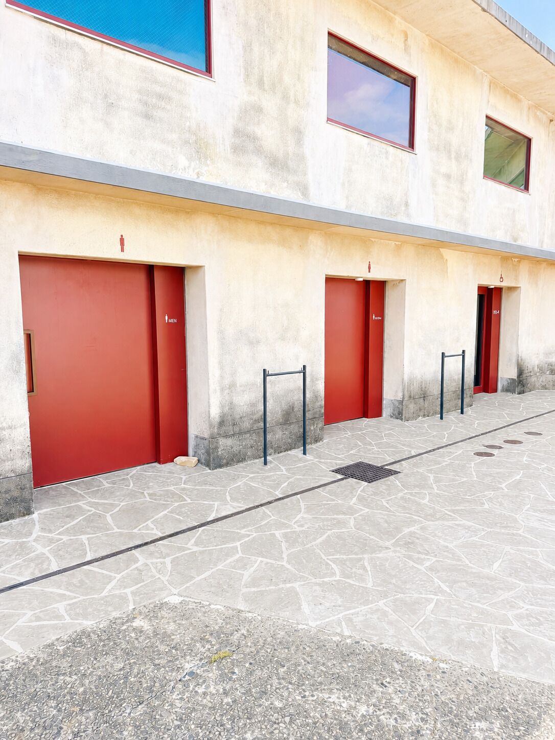 A building with red doors, each marked with a gender symbol, and a stone-paved entrance area.