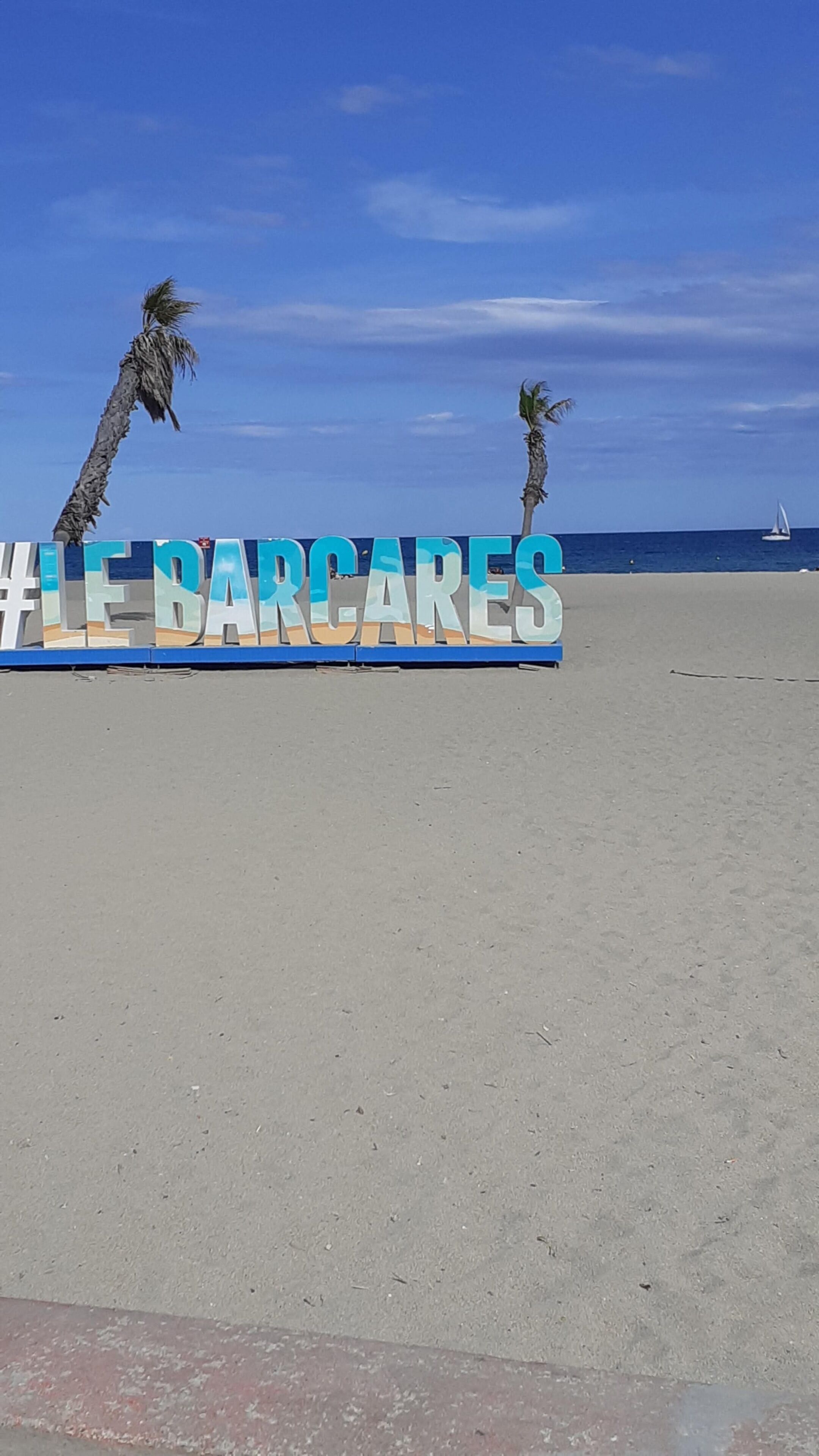 Un cartel de playa con el texto '#LE BARCARES' y palmeras al fondo.