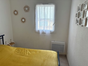 A bedroom with a bed, a window with sheer curtains, wall-mounted decor, and a radiator. - Bright village apartment with terrace and parking 50m from the sea (Le Barcarès)