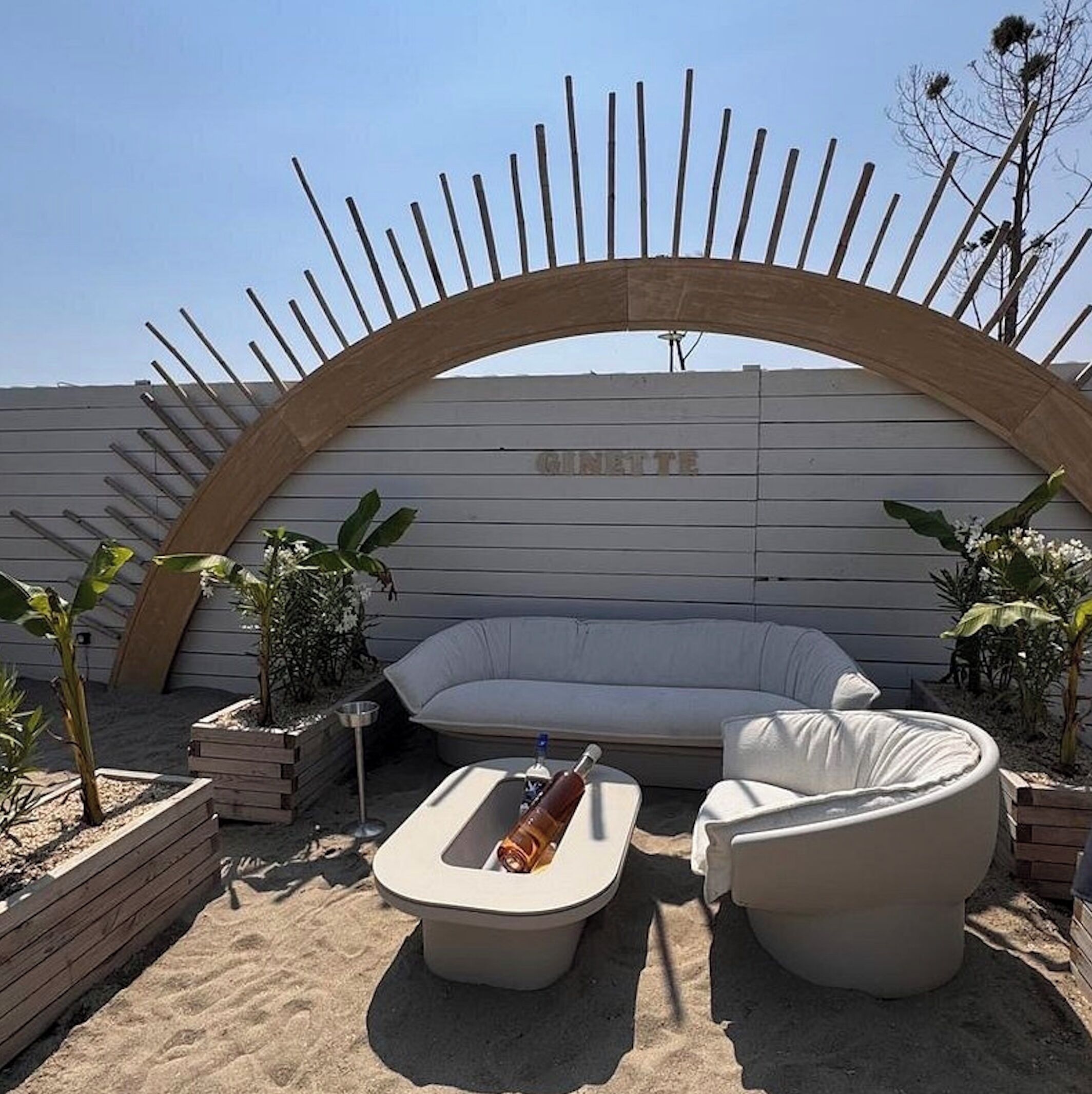 Un espace extérieur lumineux, aménagé pour se détendre, avec un canapé blanc et deux chaises blanches, une petite table, et une arche décorative en bois. Le mot « CINNette » est inscrit sur le mur situé derrière.