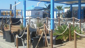 Un espace extérieur abrité, doté de tables et de chaises, d’une terrasse en bois et d’une structure bleue avec un toit.
