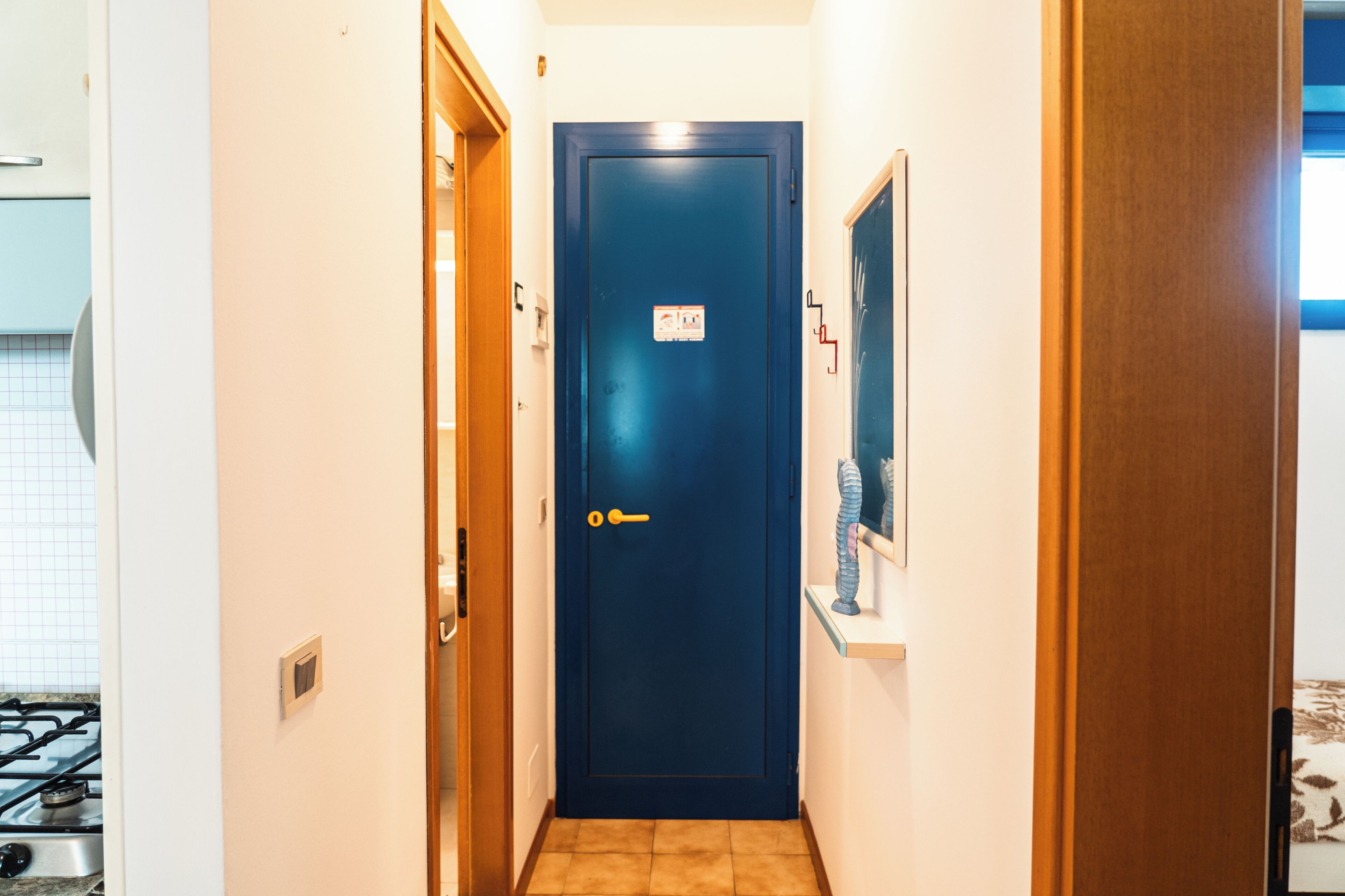 A narrow hallway with a blue door, a small shelf with a decorative item, and a tiled floor.