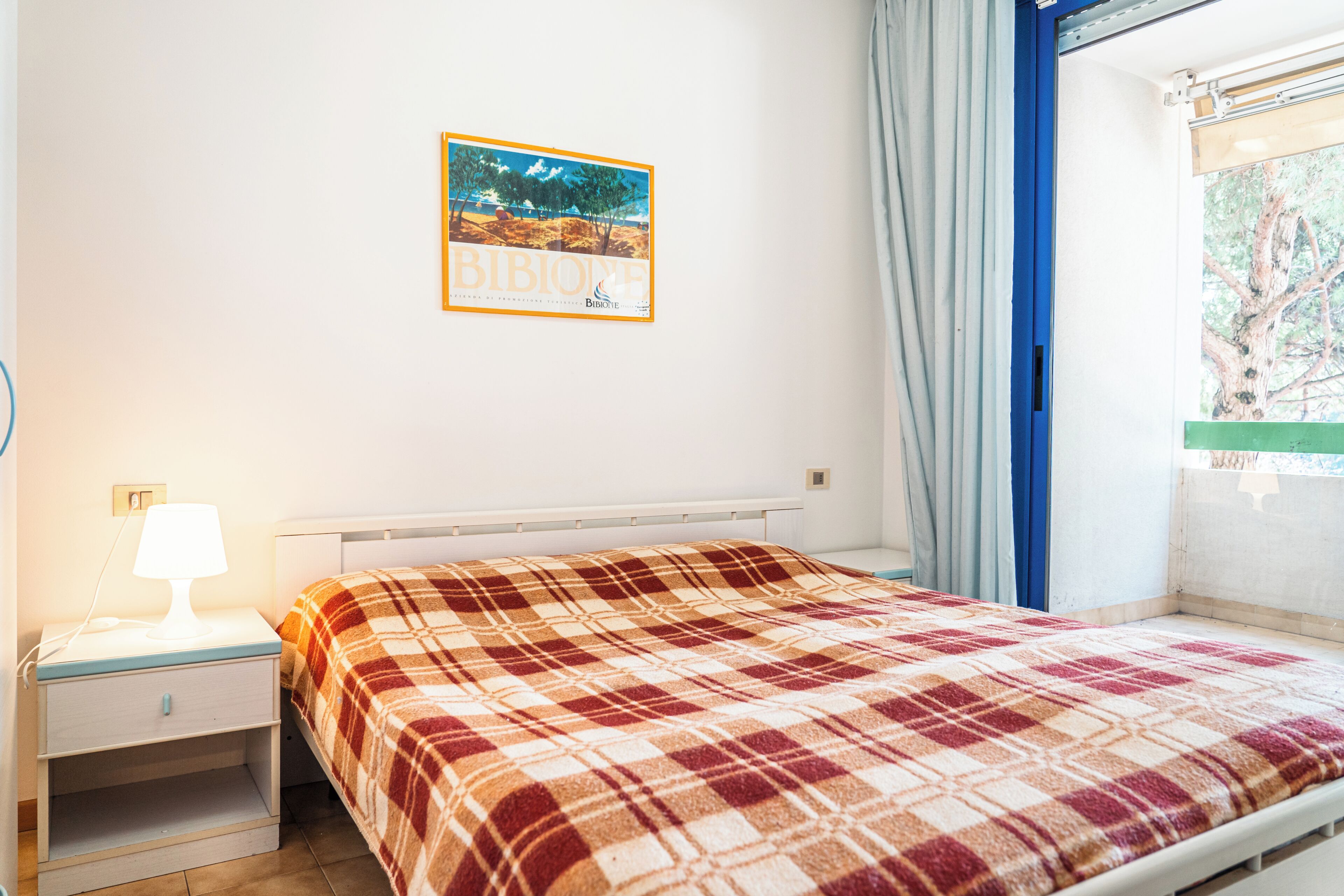 Un dormitorio con cama, mesita de noche con lámpara, un cuadro en la pared y ventana con cortinas.