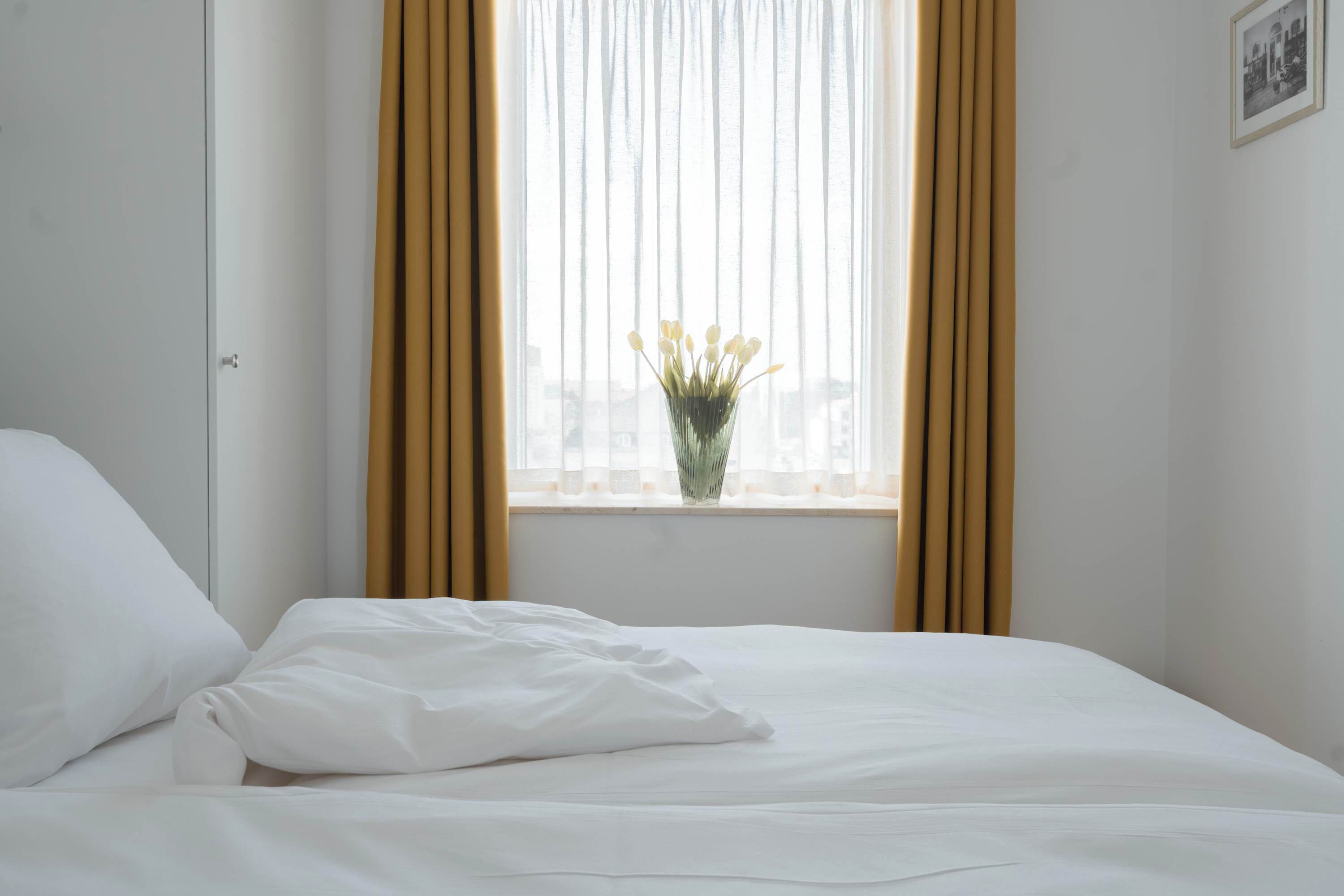 A neatly made bed with white linens, a window with sheer curtains, and a vase with flowers on the windowsill.