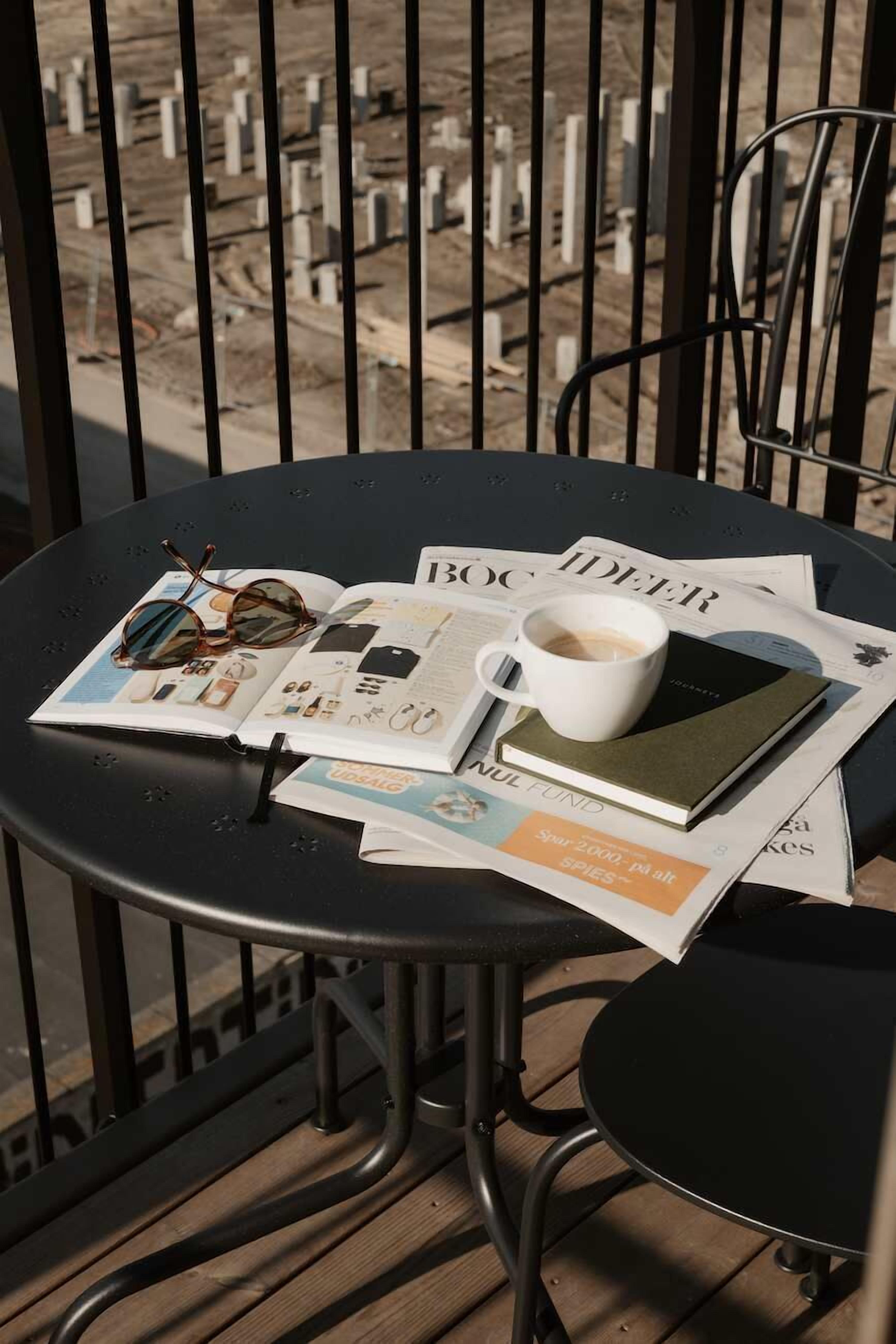 A round table with a newspaper, a book, a pair of sunglasses, and a cup of coffee on a balcony with a view of a cemetery.