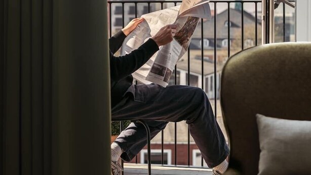 A person sitting on a balcony reading a newspaper, with a view of buildings and a church spire in the background.