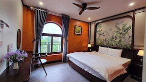 A hotel room with a bed, a desk, a chair, a ceiling fan, a window with curtains, and a tropical-themed wall mural. - Serena Tam Coc Hotel & Villa (Nam Hoa Lu)