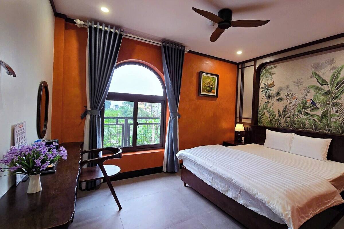 A hotel room with a bed, a desk, a chair, a ceiling fan, a window with curtains, and a tropical-themed wall mural.