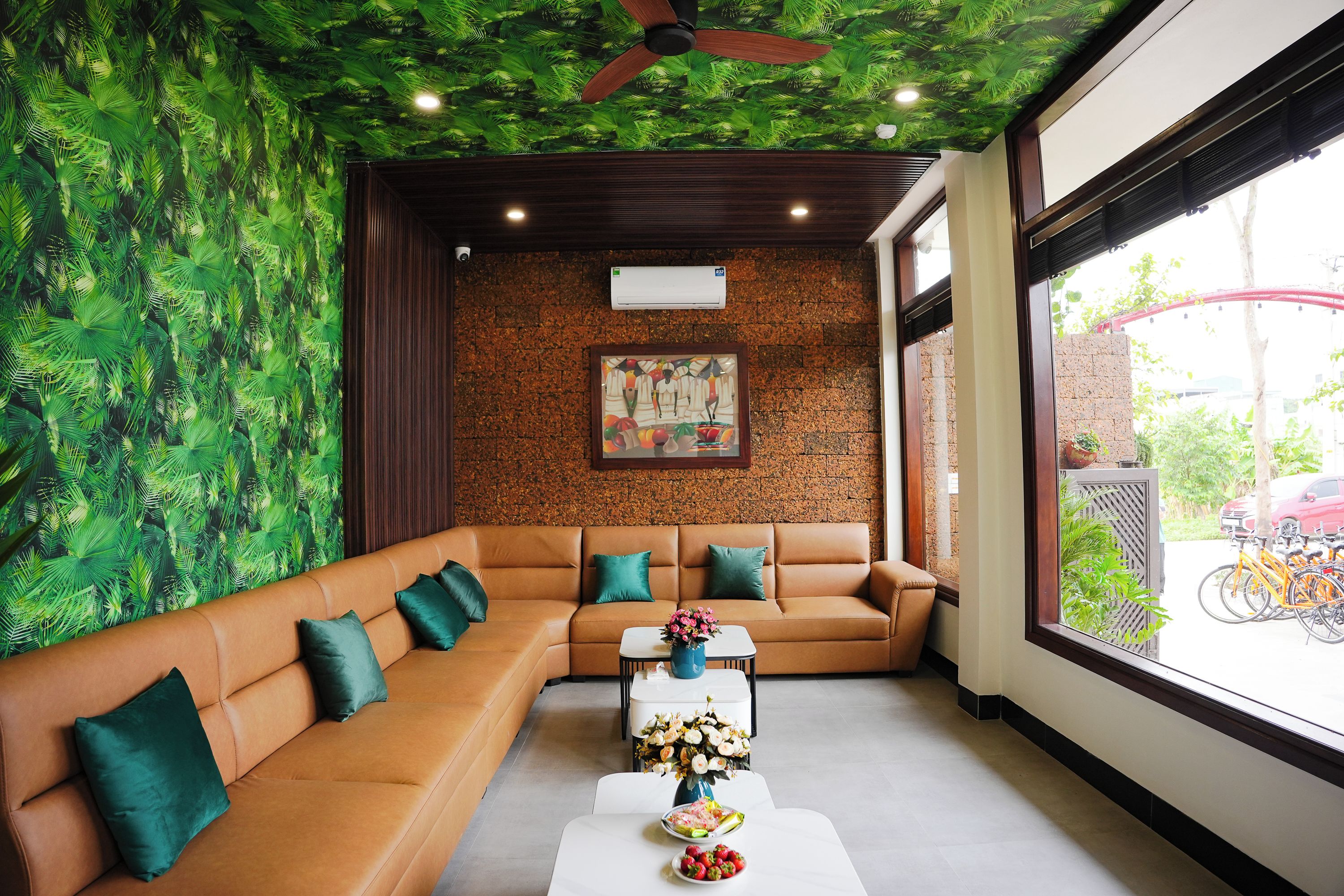 Un salon moderne avec un canapé d’angle, une table basse décorée de fleurs, une fresque murale et une vue sur des vélos à l’extérieur.
