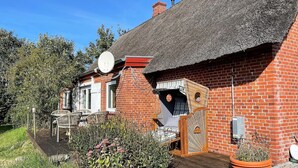 Una casa tradicional de ladrillo con techo de paja, una antena parabólica y un pequeño jardín con terraza de madera y mobiliario exterior.