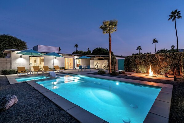 Una piscina con sillas de descanso y una chimenea, rodeada por una casa moderna con zona de patio.