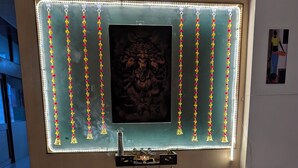 Ein Zimmer mit einem wandmontierten Fernseher, dekorativer Beleuchtung, einem kleinen Altar, Blumentöpfen und einem farbenfrohen Rangoli auf dem Boden.