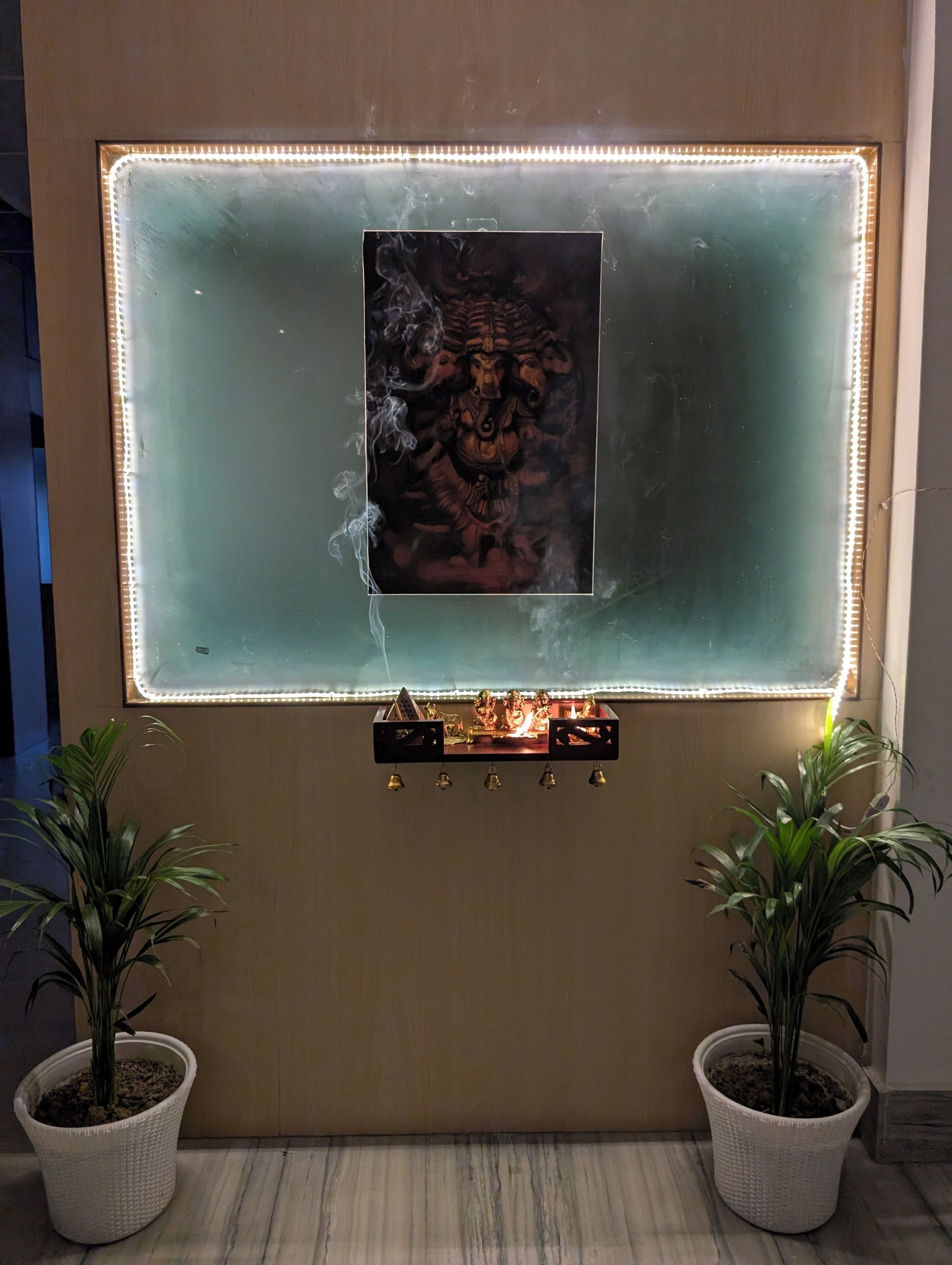 Un mur orné d’un tableau encadré, un petit autel avec des clochettes et deux plantes en pot.