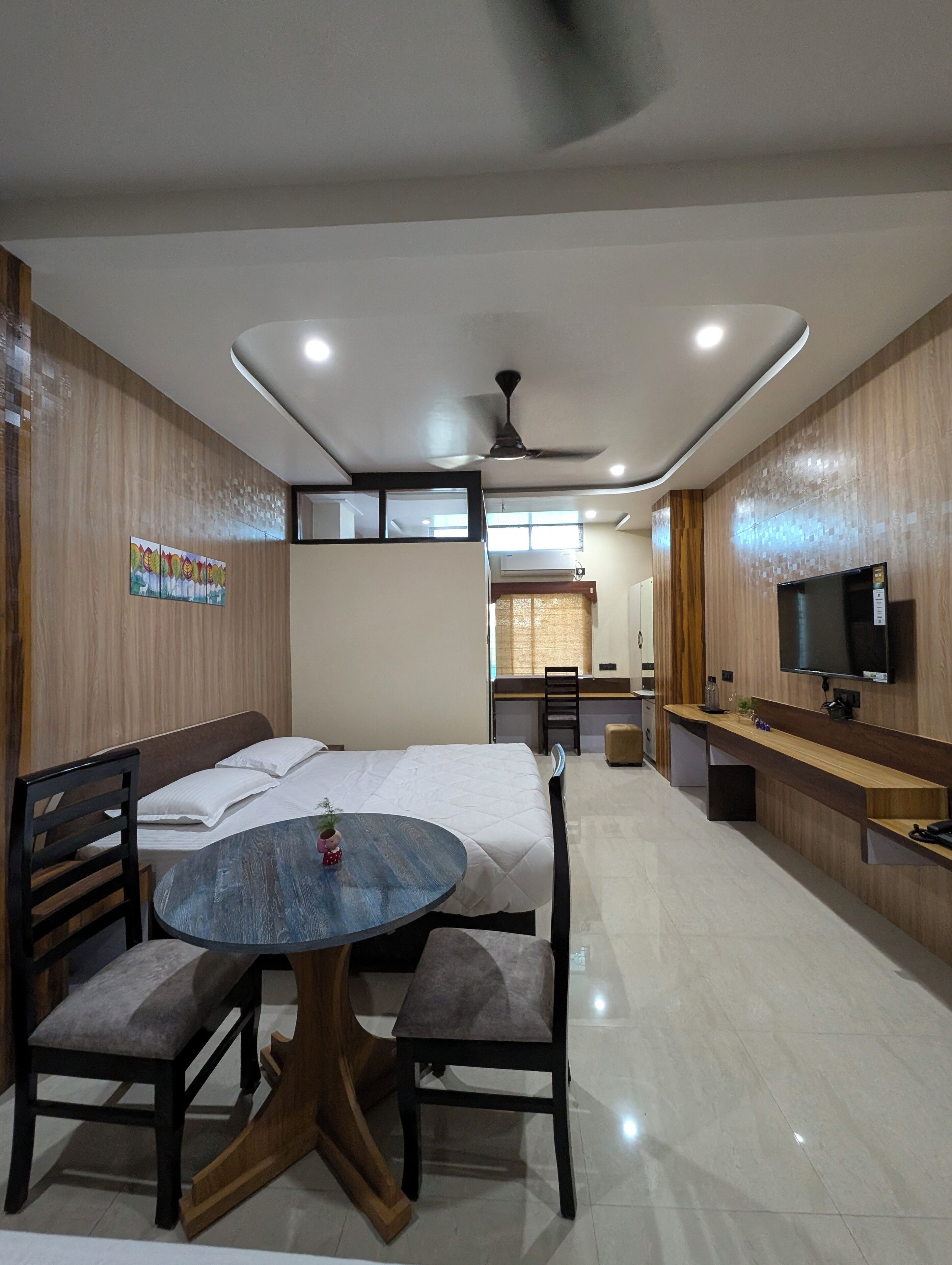 A modern hotel room with a bed, a round table with chairs, a TV, and a desk with a chair.