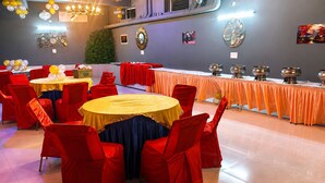 Een feestzaal met ronde tafels, rode stoelen en een lange tafel met horeca-artikelen.