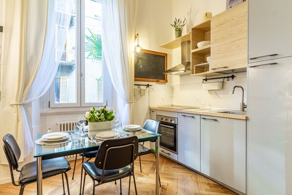 A modern kitchen with a dining table set for four, a built-in oven, and a window with sheer curtains.