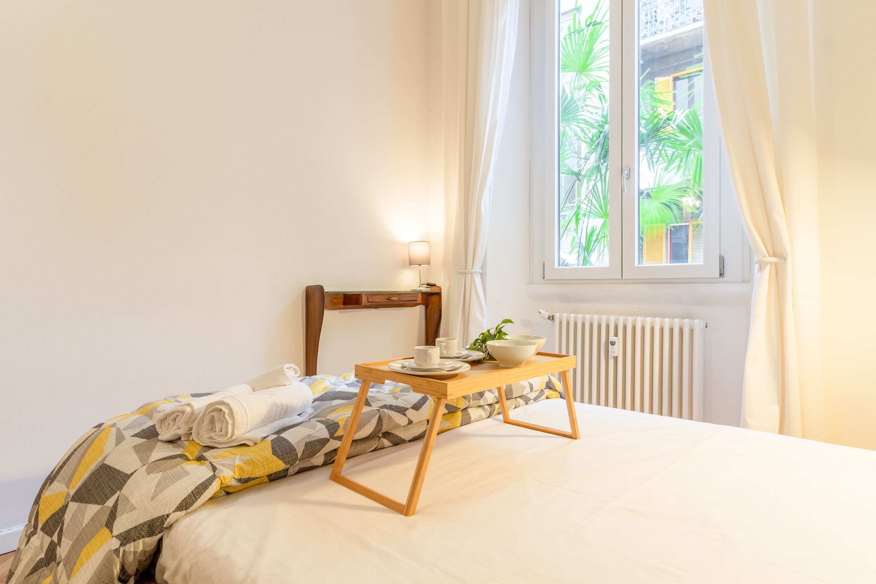 A neatly made bed with a patterned bedspread, a bedside table with a lamp, and a window with curtains.
