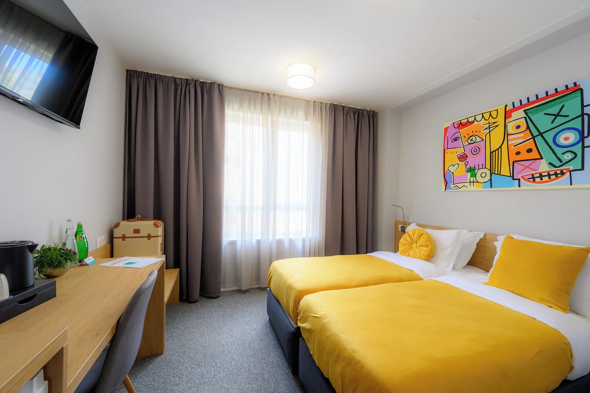 Habitación de hotel con dos camas, un escritorio, una silla, un televisor, una ventana con cortinas y un cuadro abstracto y colorido en la pared.