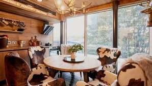 A cabin interior with a round wooden table, cowhide patterned chairs, a mounted deer head, and a large window offering a view of trees. - Student only Premium Studio at Lewisham Exchange (London)