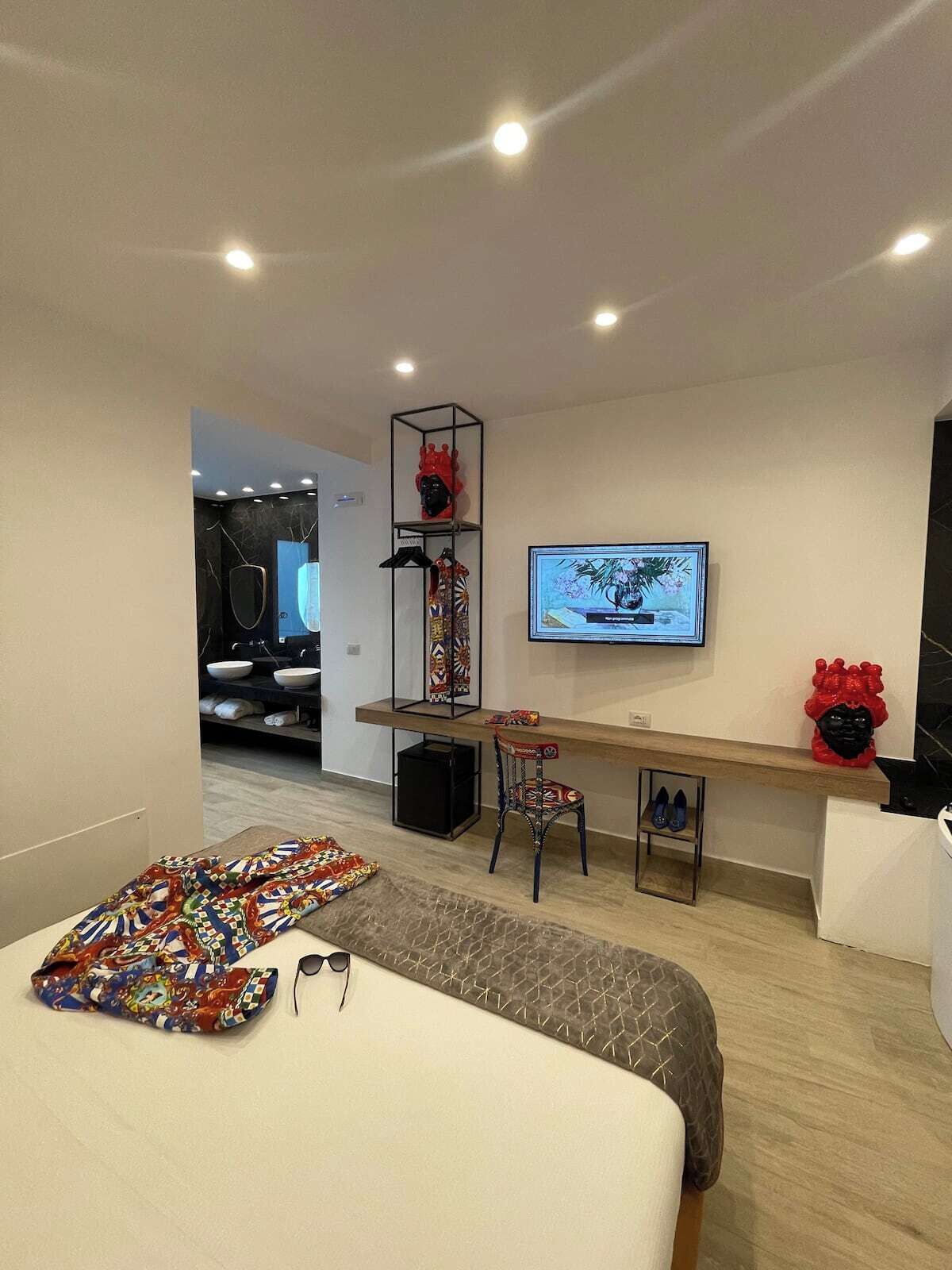 Camera d'albergo moderna con un letto, una scrivania, una sedia, una TV e un piccolo ripiano con oggetti decorativi.