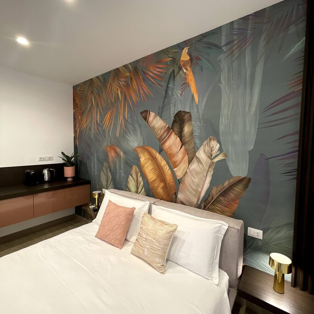 A modern bedroom with a large bed, a nightstand, a coffee maker, and a wall mural of tropical leaves.