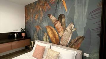 A modern bedroom with a large bed, a nightstand, a coffee maker, and a wall mural of tropical leaves.