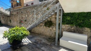 Eine Steintreppe führt zu einem Gebäude mit einem Balkon. Im Erdgeschoss steht ein Blumentopf und auf der oberen Ebene eine weiße Bank.