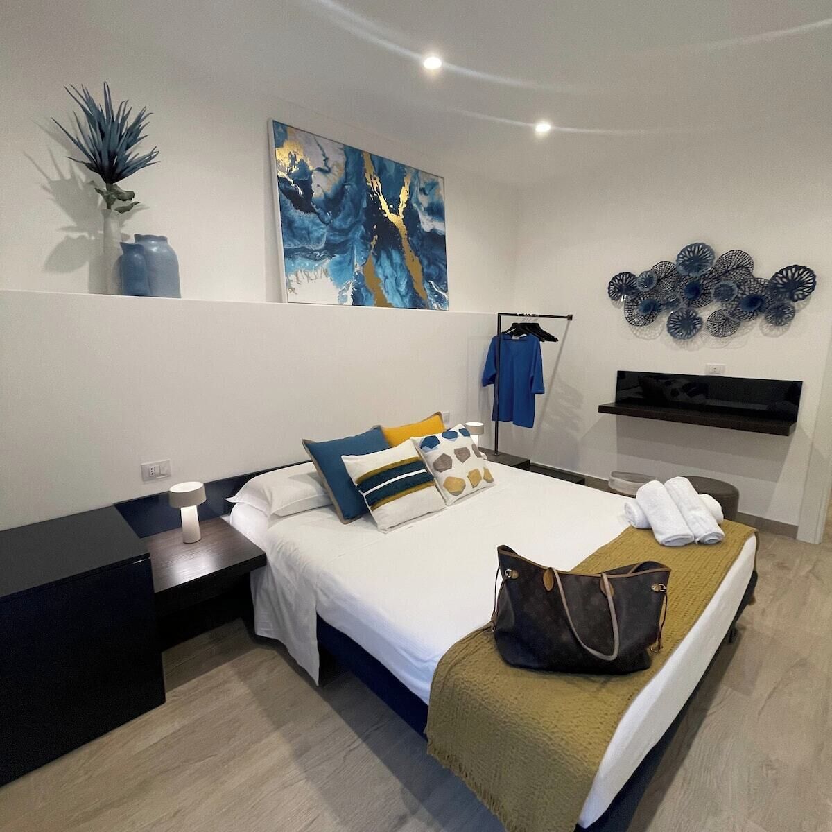 A modern hotel room with a bed, bedside tables, a nightstand, a wall-mounted TV, a decorative wall piece, and a plant.