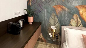 Ein schmaler Raum mit einem dunklen Holztisch, einem weißen Bett und einer Wand mit großem tropischen Blattmuster.