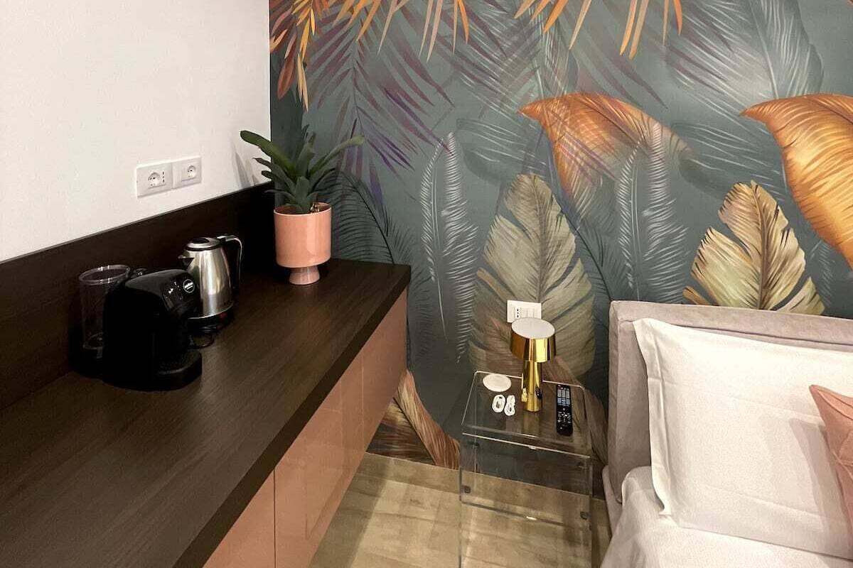 Une pièce de taille modeste, avec un bureau en bois foncé, un lit blanc et un grand mur orné d’un motif de feuilles tropicales.