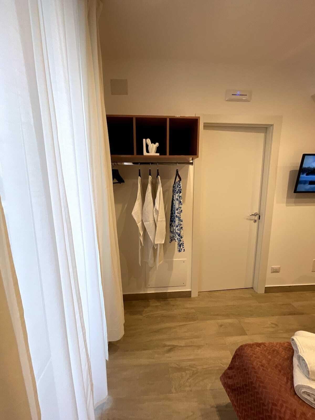 A hotel room with a wooden wardrobe, a flat-screen TV mounted on the wall, and a brown woven ottoman with a towel.