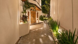 Un étroit sentier pavé mène à une porte en bois, bordée de murs blancs et de plantes vertes.