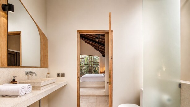 Ein Badezimmer mit Holztür zu einem Schlafzimmer, einer Toilette und einem Spiegel über dem Waschbecken.