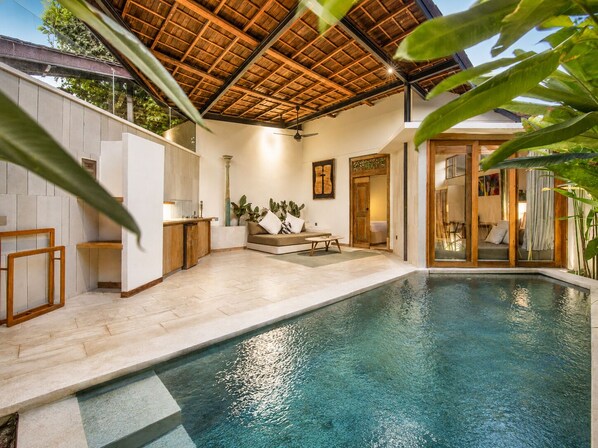 Un área interior de piscina moderna con techo de madera, una zona de estar con sofá y mesa, y una puerta de cristal que da a otra habitación.