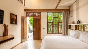A bedroom with a large bed, a wooden door, a window with a view of greenery, and a shelf with decorative items.